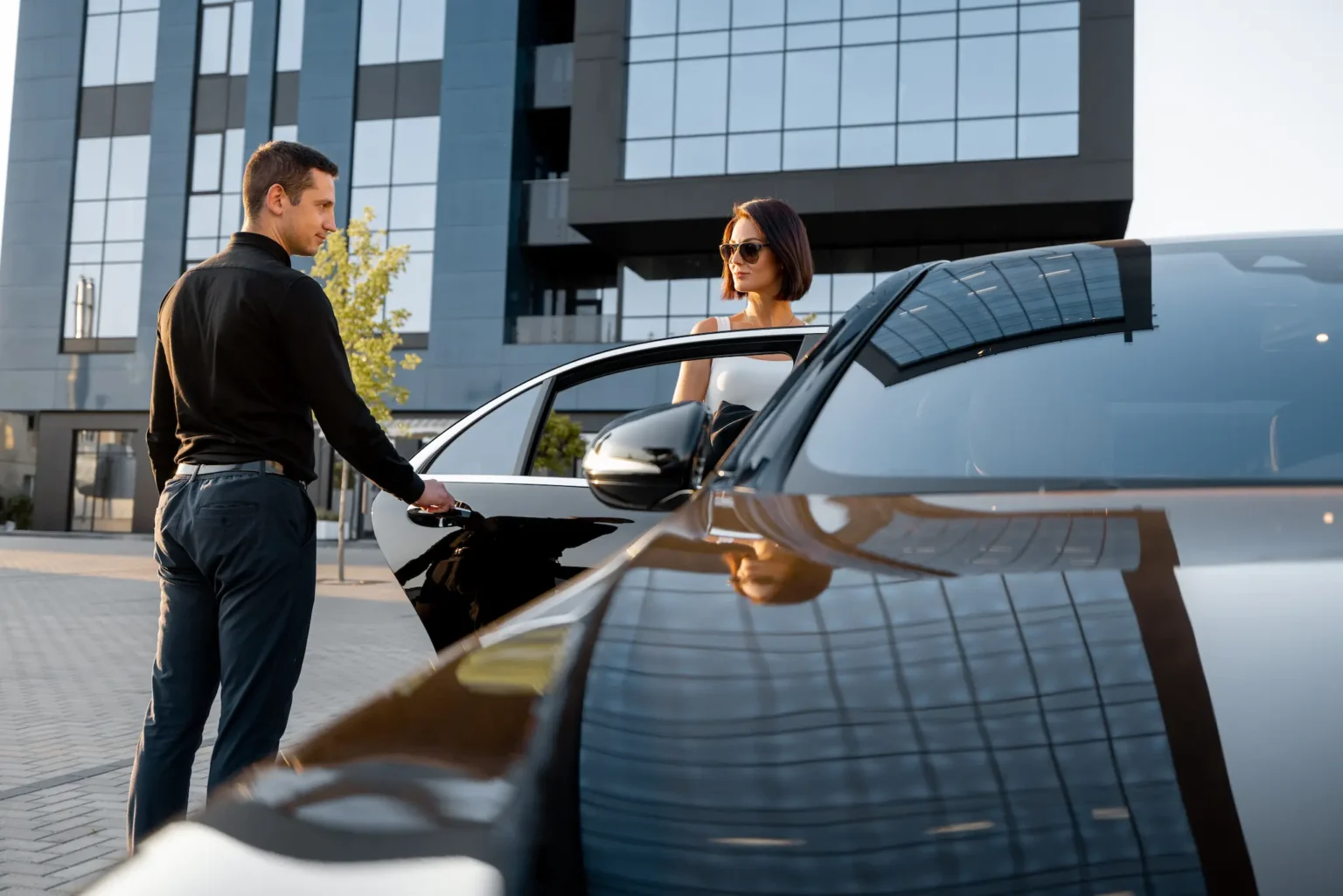 A man and woman stand beside a car, engaged in conversation, with a clear sky in the background.